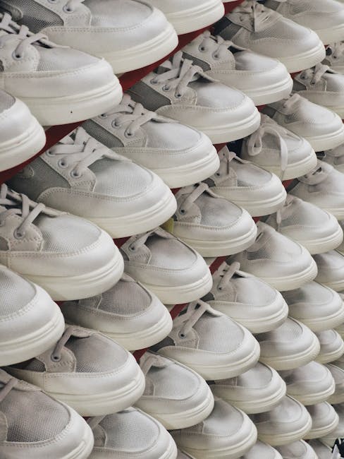 A large display of white and grey sneakers arranged in multiple rows, stacked closely together on shelves inside a retail shop or warehouse. The shoes are primarily casual trainers with white soles and laces, featuring a combination of mesh and leather-like material for the upper part. The footwear is positioned with the toes facing forward, some slightly overlapping each other, and the shoes appear to be new and unused. The background shows more shoes, indicating the space is dedicated to footwear storage or retail display, with bright lighting illuminating the area. This image exemplifies the type of footwear packing and organisation involved during home relocation or furniture transport, as coordinated by professional removals services like those offered by [COMPANY_NAME], supporting efficient packing and moving processes.