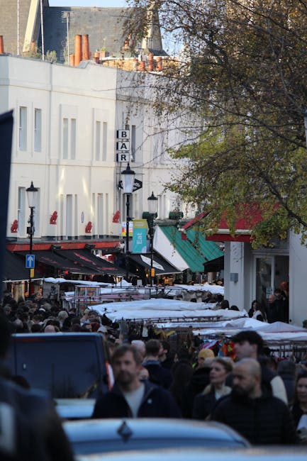 A busy street scene in a marketplace area with a large crowd of people walking along the pavement and browsing outdoor stalls covered with white and purple umbrellas. Behind the crowd, there are multi-storey buildings with shopfronts, including a bar with a vertical sign reading 'BAR' and a teal-colored awning over another storefront. The buildings are white with multiple windows, and some rooftops are visible in the background with construction materials and scaffolding. Trees with autumn-colored leaves partially obscure the view to the right, and the overall scene is illuminated by natural daylight, capturing an active environment typical of a home relocation preparation or moving process happening in an urban setting. The presence of parked vehicles and street fixtures, like street lamps and a one-way traffic sign, emphasizes the cityscape context, while the logo of Hackney Movers is referenced in relation to this moving and packing environment.