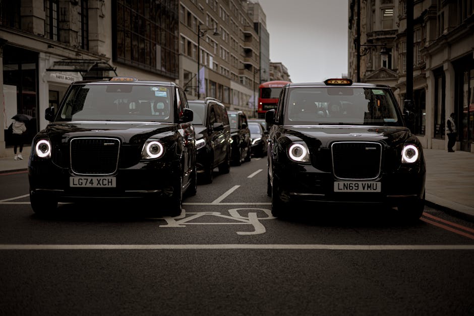 Two black London black cabs, identifiable by their distinctive shape and illuminated circular headlights, are parked side by side on a city street in an urban area. The cabs are positioned within a designated taxi zone, marked by white painted lines and the words 'TAXI' on the dark asphalt. Behind the cabs, a queue of other vehicles, including a red double-decker bus, lines the street, with additional cars visible further down. The scene is set in a dense area with multi-storey buildings lining both sides of the street, featuring a mix of modern and historic architecture, windows, and storefronts. Pedestrians walk along the sidewalk, some carrying bags, and a few are near the buildings. Overcast skies cast diffuse lighting on the scene, and the overall atmosphere reflects busy city traffic, related to urban transportation and parking, relevant to house removals or moving services like those offered by [COMPANY_NAME] at hackneymovers.co.uk, especially in the context of moving or relocation logistics.