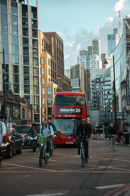 A red double-decker bus displaying route number 26 and the destination 'Hackney Wick' is approaching a busy urban intersection in central Hackney, surrounded by modern glass and brick office buildings and residential apartments. Several pedestrians and cyclists, some wearing backpacks, are crossing the street and waiting at the traffic lights. The scene captures the dense city environment with tall skyscrapers and a partly cloudy sky overhead. This image relates to house removals and moving services, illustrating urban transport and travel logistics in the Hackney area, which Hackney Movers often support for home relocations and furniture transport in Dalston Kingsland and surrounding streets.