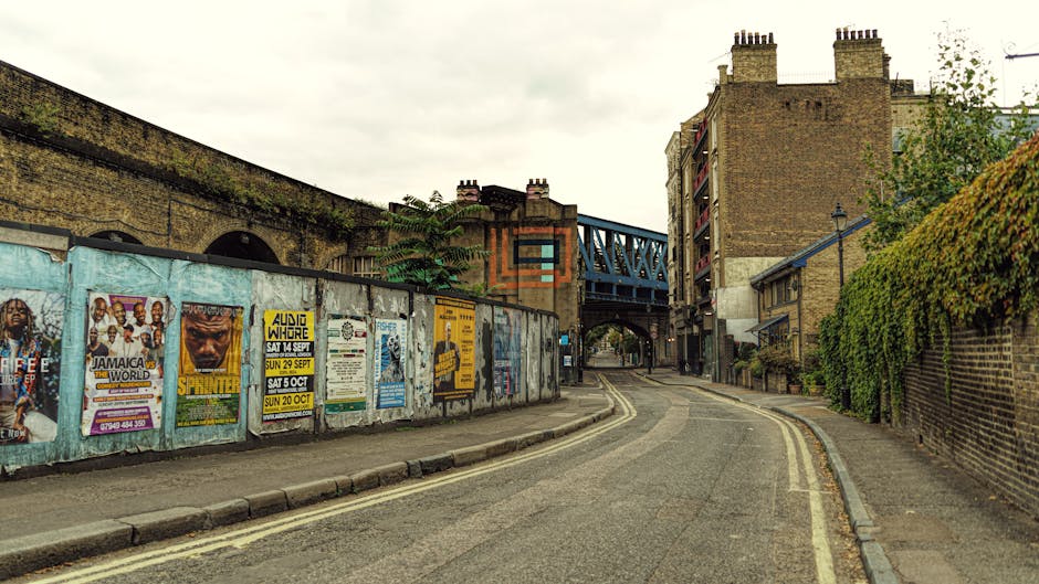 A narrow street in Dalston Kingsland during a home relocation scene, showing a loading area near the pavement with cardboard boxes and wrapped furniture, likely prepared for transportation by Hackney Movers' removals team. The street features a brick wall on one side decorated with posters and a green hedge on the other, with residential buildings and a blue bridge structure crossing above in the background. The scene suggests a professional furniture transport and packing process, with the loading zone set up to facilitate the moving of household items through tight, urban streets. The overcast sky provides diffuse lighting, and the environment indicates an active residential area suitable for detailed house removals, including packing, loading, and transport preparation.