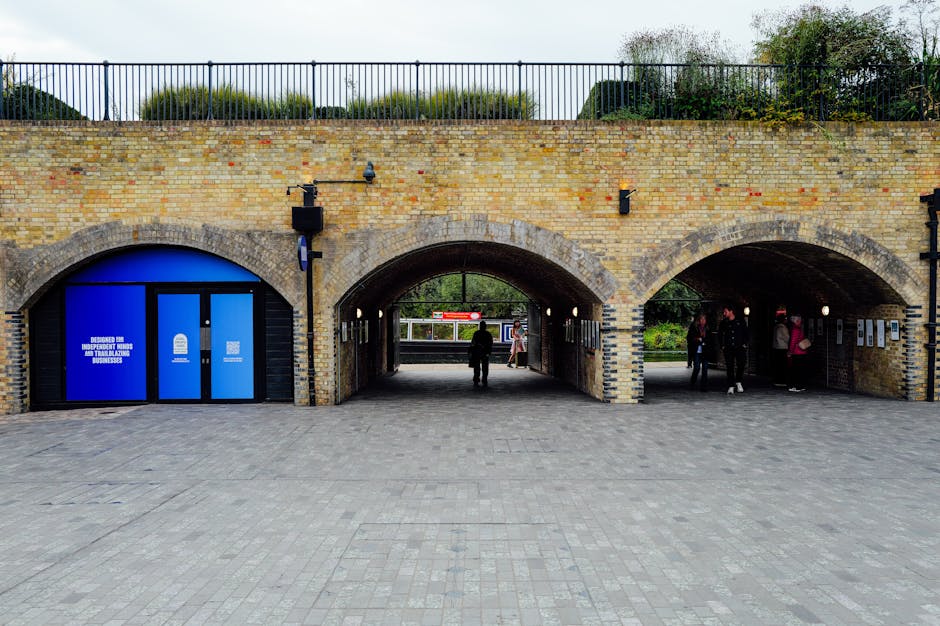The image shows a brick railway station underpass with three archways leading to different areas outside. The left arch has a blue door with White logos and text indicating accessibility for independent businesses, suggesting a commercial or office entrance. The middle arch is open, revealing a paved area with a person walking through, and beyond, the platform of Hackney Wick station with a visible train and a few passengers. The right arch contains several people gathered, some standing and others walking, possibly waiting or preparing for a home relocation or furniture transport process. The brickwork is light-colored with darker brick accents around the arches, and the area is lit by mounted black-wash lights. Above the structure, a black metal railing runs along the top of the brick wall with greenery visible behind it. The scene is captured during daylight with overcast sky, providing natural light that highlights the textures of the bricks, paving stones, and surroundings, illustrating a typical scene of moving logistics involving loading exterior areas connected to a transport station.
