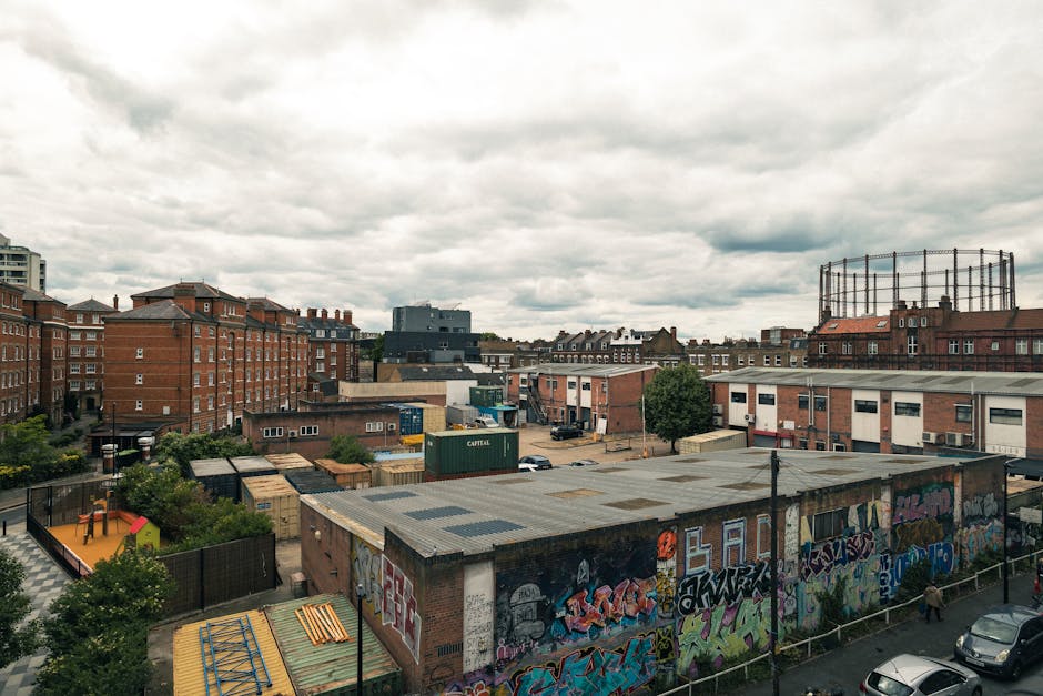 A wide view of an urban area showing multiple brick residential buildings and commercial structures under a cloudy sky, with a rooftop parking area and a graffiti-covered warehouse in the foreground. The scene includes a fenced playground with a slide to the left, loads of cardboard boxes and packing materials stacked on the roof of a building, and a parking lot with several parked cars. Visible are loading and transport activities related to home relocation, with a few moving boxes and packing supplies scattered around. The image captures a typical city environment during daytime with overcast weather, emphasizing the logistical process of furniture transport and packing involved in house removals, as seen in professional services such as those offered by [COMPANY_NAME], situated within the context of a Mare Street Hackney house removals guide.