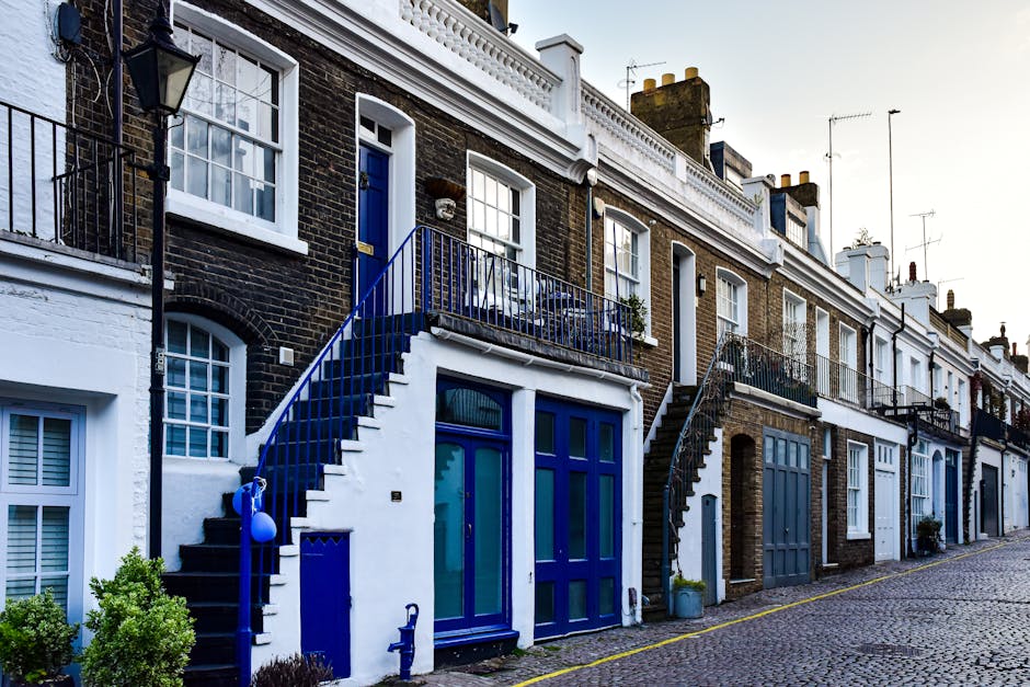A row of terraced houses on a cobbled street in Hackney, with each property featuring a staircase leading from the pavement to the front door. The houses have traditional brick exteriors with white decorative trims, large windows, and black or blue-painted doors. Some staircases have metal railings painted in blue, with a few decorated with potted plants or small outdoor items. The street scene includes various bicycles and potted plants near entrances. This exterior view depicts a typical London residential area suitable for house removals, illustrating the environment where furniture transport and home relocation activities, such as carrying, loading, and packing, may take place by professional removals services like Hackney Movers, to facilitate moving and moving logistics.
