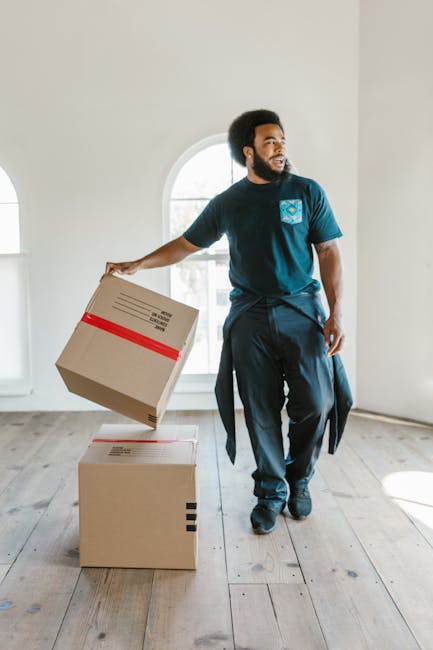 A man with a beard and curly hair, dressed in a navy blue t-shirt and matching pants, is inside a well-lit room with white walls and a large arched window. He is holding a cardboard box with red tape in his right hand, preparing to place it on top of another cardboard box already on the wooden floor. Behind him, the room appears to be empty with no furniture visible, indicating a home relocation or packing process. The natural light from the window highlights the moving activity, suggesting an organized packing and loading process carried out by professional removals, possibly associated with Hackney movers for same-day relocation services. The scene reflects a professional approach to furniture transport and packing during quick, efficient home moves, with the focus on careful handling of cardboard boxes and planning for effective space management during interior packing and moving procedures.