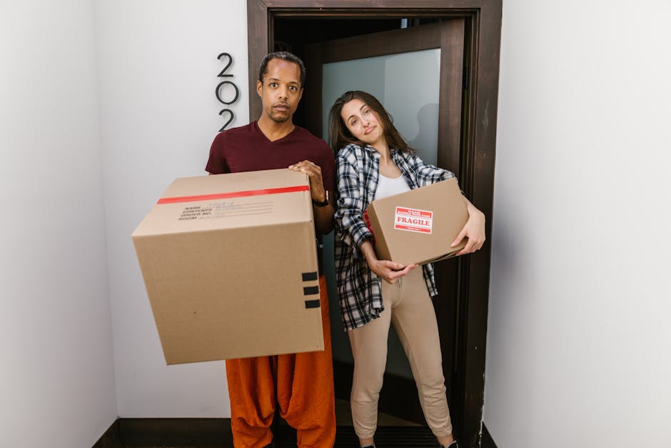 A man and a woman stand in a narrow hallway beside an open doorway, each holding a cardboard box for a home relocation. The man is wearing an orange pair of trousers and a maroon shirt, holding a large box in front of him, while the woman is dressed in light beige pants and a checkered shirt, holding a medium-sized box labeled 'Fragile' close to her chest. Behind them, a dark wooden door frame surrounds a glass panel, and the area is illuminated by natural or ambient lighting. The scene suggests an active loading process typical of furniture transport and packing activities involved in domestic removals, with the focus on careful handling of packed items and spatial arrangement within the property for efficient home relocation, as performed by [COMPANY_NAME] or similar removals services in Hackney.